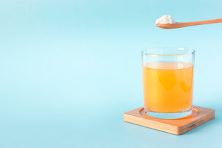 A spoon of collagen hovering over a glass of juice against a bright blue background