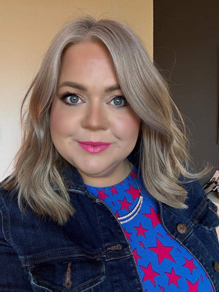 A person with shoulder-length blonde hair is shown smiling at the camera. They are wearing a blue top with red stars and a denim jacket
