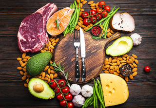 A wooden cutting board with a knife and fork is surrounded by various foods, all placed on a dark wooden surface.