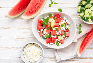 A white bowl with watermelon and feta salad. The bowl is surrounded by watermelon slices, a bowl of cucumber and a bowl of feta. All item are on a white wooden tabletop 