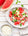 A white bowl with watermelon and feta salad. The bowl is surrounded by watermelon slices, a bowl of cucumber and a bowl of feta. All item are on a white wooden tabletop 