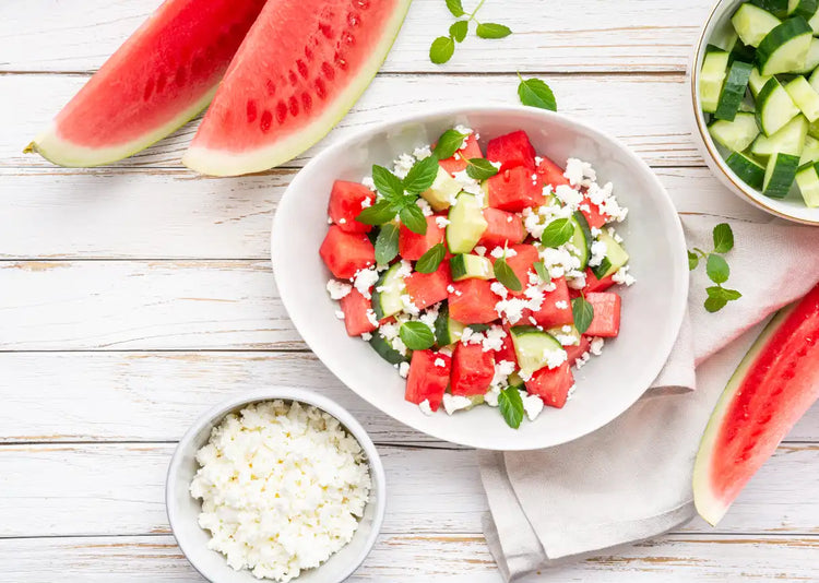A white bowl with watermelon and feta salad. The bowl is surrounded by watermelon slices, a bowl of cucumber and a bowl of feta. All item are on a white wooden tabletop 