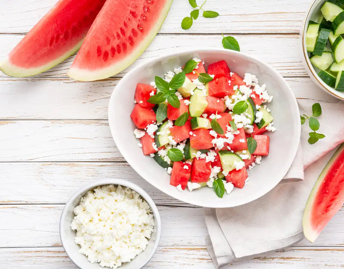 A white bowl with watermelon and feta salad. The bowl is surrounded by watermelon slices, a bowl of cucumber and a bowl of feta. All item are on a white wooden tabletop 
