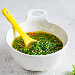 A white ceramic bowl filled with vibrant green coriander sauce sits on a light gray surface. A yellow-handled spoon rests inside the bowl, and the sauce is speckled with red chili flakes and herbs.