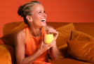 Willpowders founder Davinia Taylor laughing with a yellow drink, sitting on an orange sofa on an orange background 