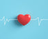 A red heart shape is centered on a light blue background. A white electrocardiogram (EKG) line runs horizontally across the image, passing through the heart. The EKG line has peaks and valleys representing a heartbeat.