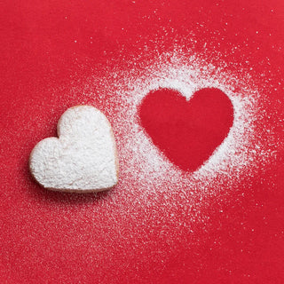 A heart-shaped cookie dusted with powdered sugar lies on a red surface. Next to it is an empty heart-shaped outline created by the dusted sugar, contrasting sharply against the red background.