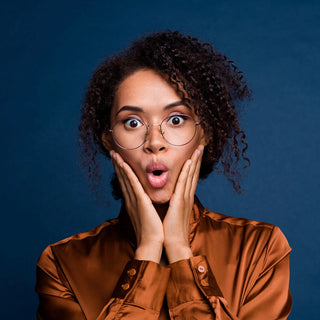A person with curly hair and wearing glasses is looking surprised, with wide eyes and an open mouth, and both hands placed on their cheeks, dressed in a shiny brown shirt
