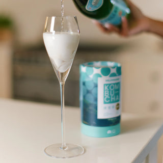 Person pouring a drink into a glass with a can of kombucha in the background.