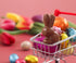 A miniature shopping cart filled with various colorful foil-wrapped chocolates and a chocolate bunny sits on a pink surface