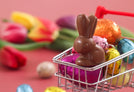 A miniature shopping cart filled with various colorful foil-wrapped chocolates and a chocolate bunny sits on a pink surface