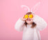 A child wearing a fluffy white costume with pink bunny ears is standing against a pink background