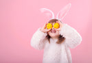 A child wearing a fluffy white costume with pink bunny ears is standing against a pink background