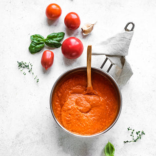 A pot of red tomato sauce with a wooden spoon , surrounded by tomatoes, garlic, basil & thyme sprigs