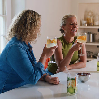 Two women sitting at a table with drinks, smiling and clinking glasses.