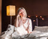 A person with short blonde hair sits on a bed with white sheets, holding their head with a tired expression