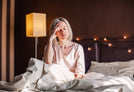 A person with short blonde hair sits on a bed with white sheets, holding their head with a tired expression