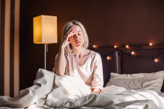 A person with short blonde hair sits on a bed with white sheets, holding their head with a tired expression