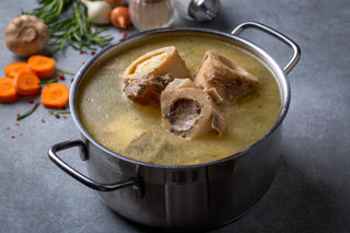 A stainless steel pot filled with bone broth, showcasing large marrow bones. The pot is placed on a grey countertop, surrounded by ingredients like sliced carrots, garlic, onions, and rosemary in the background, suggesting a fresh and wholesome preparation of the broth.