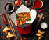 A white Chinese takeaway box filled with fried rice, spring rolls, and sautéed vegetables, placed on a dark slate surface. Wooden chopsticks rest on the side, and scattered grains of rice and sliced vegetables surround the box, enhancing the appetizing presentation.