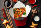 A white Chinese takeaway box filled with fried rice, spring rolls, and sautéed vegetables, placed on a dark slate surface. Wooden chopsticks rest on the side, and scattered grains of rice and sliced vegetables surround the box, enhancing the appetizing presentation.