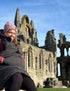 A person wearing a pink hat and black jacket sits on a stone wall in front of the ruins of an ancient stone abbey.