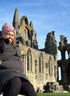 A person wearing a pink hat and black jacket sits on a stone wall in front of the ruins of an ancient stone abbey.