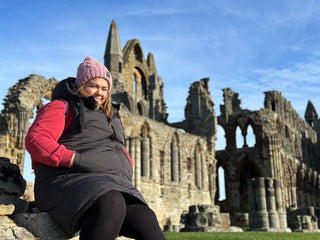 A person wearing a pink hat and black jacket sits on a stone wall in front of the ruins of an ancient stone abbey.