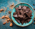 A turquoise bowl of dark chocolate on a blue countertop surrounded by cocoa beans
