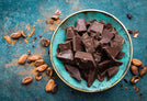 A turquoise bowl of dark chocolate on a blue countertop surrounded by cocoa beans