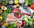 A variety of fresh foods are displayed on a dark surface, including raw meat, fish, chicken, fruits, vegetables, nuts, seeds, eggs, olive oil, blueberries, avocado, pomegranate, lemon, and assorted greens.