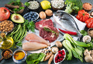 A variety of fresh foods are displayed on a dark surface, including raw meat, fish, chicken, fruits, vegetables, nuts, seeds, eggs, olive oil, blueberries, avocado, pomegranate, lemon, and assorted greens.
