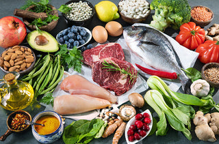 A variety of fresh foods are displayed on a dark surface, including raw meat, fish, chicken, fruits, vegetables, nuts, seeds, eggs, olive oil, blueberries, avocado, pomegranate, lemon, and assorted greens.