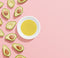 Halved avocados on the left of a pink background. A white bowl filled with yellow oil