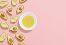 Halved avocados on the left of a pink background. A white bowl filled with yellow oil