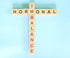 Wooden letter tiles spell "HORMONAL" horizontally and "IMBALANCE" vertically on a light blue background.