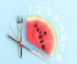 A watermelon on a light blue background, resembling the section of a clock. Knife and fork represent the clock hands