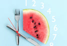A watermelon on a light blue background, resembling the section of a clock. Knife and fork represent the clock hands