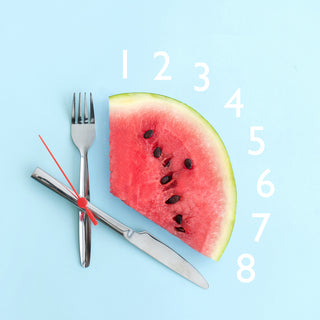 A watermelon on a light blue background, resembling the section of a clock. Knife and fork represent the clock hands