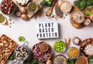 A variety of plant-based protein sources are displayed around a white box "PLANT BASED PROTEIN", arranged on a table.