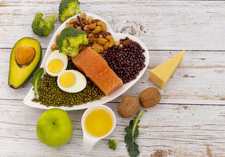A selection of healthy foods on a white wooden table, salmon, boiled eggs, nuts, seeds arranged on a heart-shaped dish.