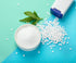 A white bowl filled with sugar on a blue and green background. Next to multiple sweeteners and stevia leaves