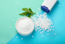 A white bowl filled with sugar on a blue and green background. Next to multiple sweeteners and stevia leaves