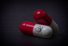 Two red and white capsules, one with a skull and crossbones symbol, lie on a dark surface. The symbol indicates a warning, potentially suggesting that the capsules are harmful or toxic.